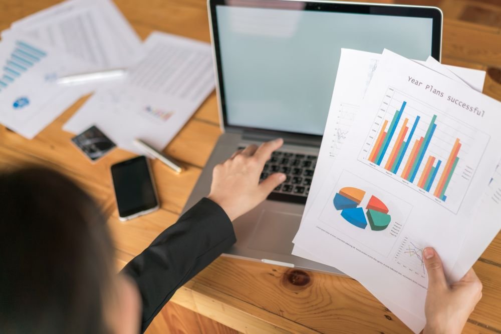 muškarac pokazuje na laptop dok u desnoj ruci drži statistiku na papirima